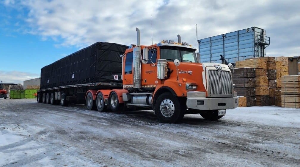 Orange flatbed truck secured with heavy duty tarp straps and a black Verduyn tarp system, showing proper cargo securement setup for a tarp straps buyers guide